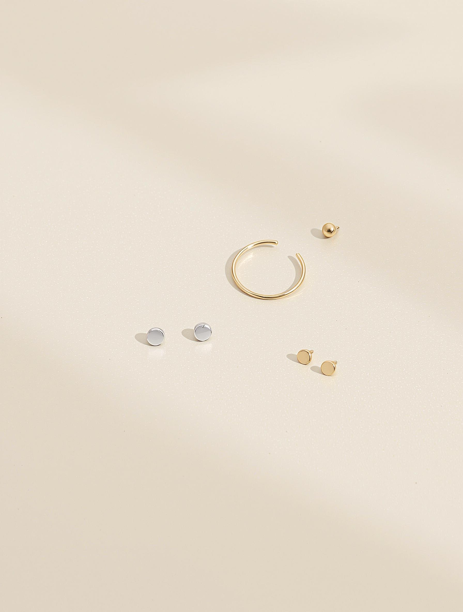 Trois paires de boucles d’oreilles délicates sur fond neutre : puces en argent 925, mini créoles plaqué or, ear cuff minimaliste gold filled.